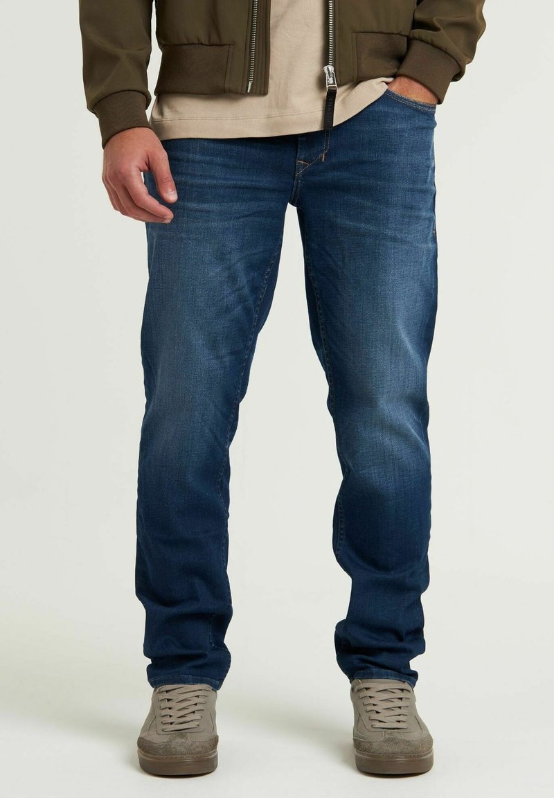 Jean en denim bleu à coupe droite, avec une légère décoloration et un classique design à cinq poches, associé à des baskets grises et un t-shirt marron clair.