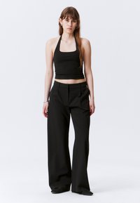 Young woman with long brown hair wearing a black halter crop top, wide-leg black trousers, and black shoes against a plain white background.