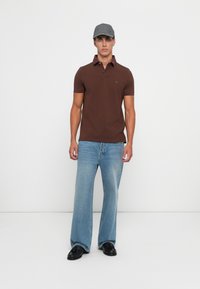 Ung mann iført grå caps, brun kortermet polo, lyseblå vide jeans og svarte sko stående mot en hvit bakgrunn.