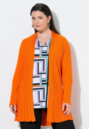 Kobieta w jasnopomarańczowym prążkowanym kardiganie nałożonym na bluzkę z geometrycznym wzorem oraz czarnych spodniach, stojąca na tle białej ściany.