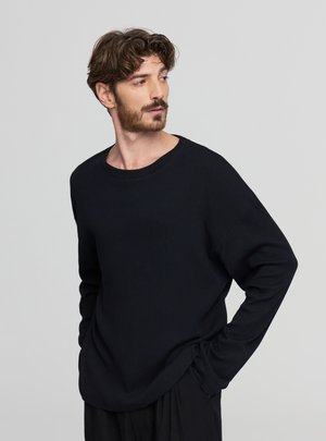 Uomo con capelli castani e barba che indossa un maglione nero a maniche lunghe, che guarda di lato con le mani nelle tasche, su uno sfondo grigio uniforme.