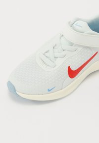 Zapatilla deportiva de color gris claro con parte superior de malla, logo de Nike rojo, detalles azules y una correa de Velcro. Punta redondeada y suela acolchada.