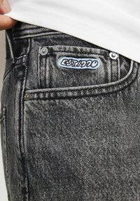 Harmaat farkut, joiden taskussa on pieni etiketti, jossa lukee "EST. 1990", ommeltuna taskun yläpuolelle, osittain peitettynä valkoisella paidalla.