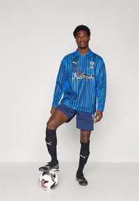 Camisola de futebol de mangas compridas com riscas azuis e texto dourado, calções azul-marinho, meias pretas até ao joelho e chuteiras pretas, em cima de uma bola.