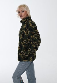 Jacke in felpa verde mimetica con collo alto, chiusura a zip e texture morbida. Abbinata a jeans azzurri chiari con ricami colorati.