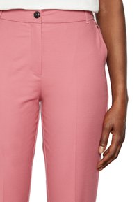 Rosa byxor med en slät yta, med en knapp i fronten, sidofickor och sydda sömmar, visade med en hand som vilar på tyget.