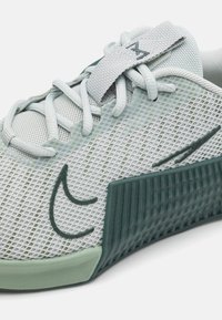 Világosszürke sportcipő hálós felsőrésszel, zöld texturált talp, fűzős kialakítással és egy fekete logóval ellátott textil pánttal a tetején.