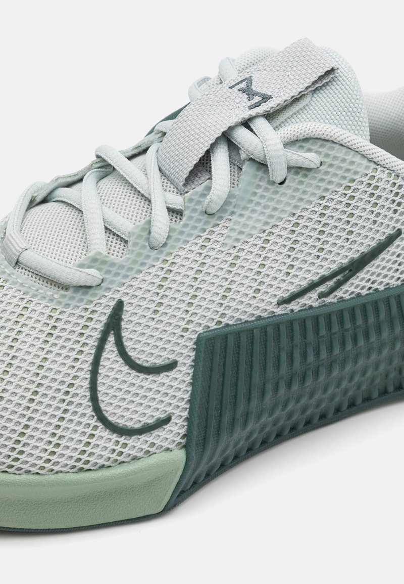 Zapatilla deportiva gris claro con parte superior de malla, suela texturizada verde, diseño con cordones y una correa de tela con un logo negro en la parte superior.