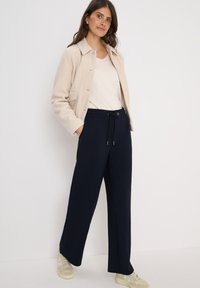 Beige fleecejack met kraag, gecombineerd met marineblauwe broek met elastische taille en lichtgekleurde sneakers. Model staat tegen een witte achtergrond.