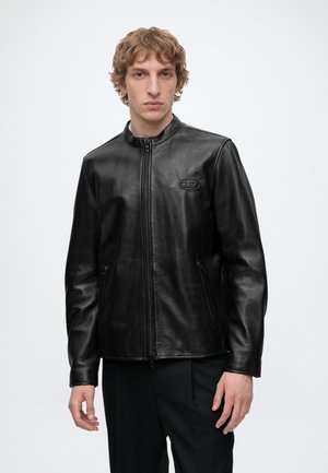 Junger Mann mit kurzen lockigen Haaren, der eine schwarze Lederjacke mit Reißverschluss und kleinem Logo auf der Brust sowie schwarze Hose trägt, steht vor weißem Hintergrund.