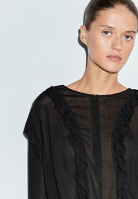 Svart genomskinlig topp med en dragkedja framtill och volangdetalj längs halsringningen. Enkelt mönster med en slät textur.