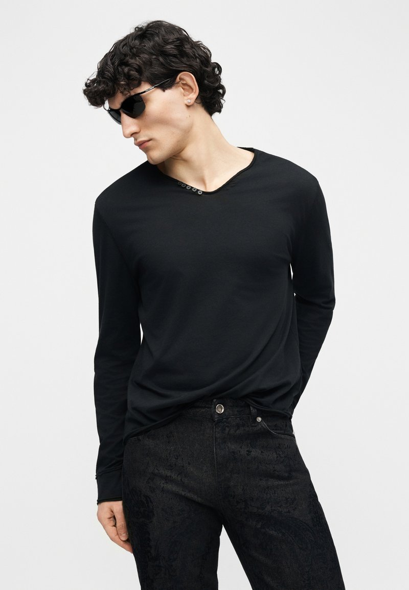 Uomo con capelli ricci che indossa una camicia nera a maniche lunghe con bottoni vicino al colletto e pantaloni neri con motivi, che guarda di lato con occhiali da sole.