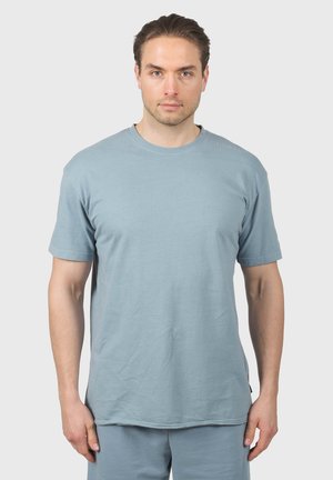 Man met een lichte huid en bruin haar draagt een effen lichtblauw T-shirt met korte mouwen en bijpassende broek tegen een lichtgrijze achtergrond.