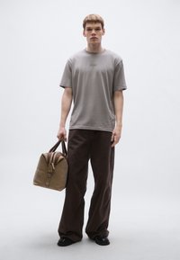 Szürke rövid ujjú póló hímzett logóval, laza barna nadrággal párosítva. A modell egy barna részletekkel díszített világosbarna hétvégi táskát tart.