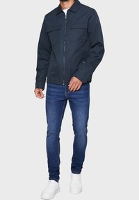 Mørk navy jakke lavet af letvægtsstof, med to brystlommer, lynlås foran og krave. Kombineret med tætsiddende blå jeans og hvide sneakers.