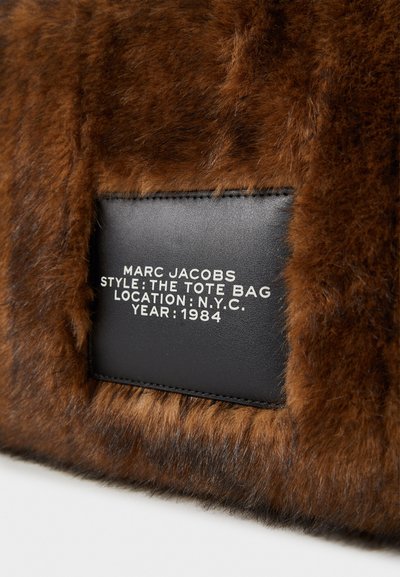 Hnědá kabelka z umělé kožešiny s černým koženým štítkem. Štítek obsahuje bílý text: "MARC JACOBS, STYL: TOTE BAG, MÍSTO: N.Y.C., ROK: 1984."