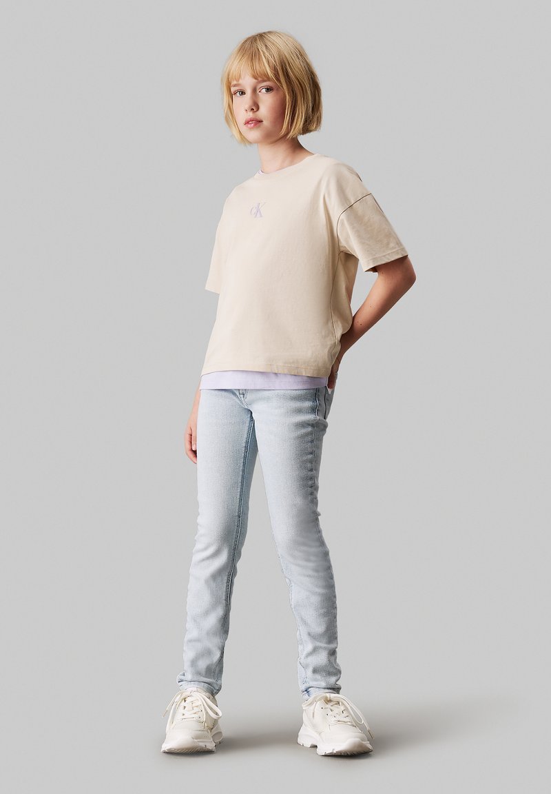 Beige pamut póló rövid ujjú, egy kis lila logóval; világoskék skinny farmernadrággal és fehér sportcipővel párosítva.