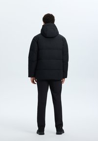 Zwarte gewatteerde jas met capuchon, voorzien van rechthoekige stiknaden, lange mouwen en een rechte snit. Gekleed met zwarte broek en schoenen.