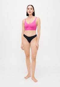 Kvinne som står barføtt iført en lys rosa blonde-bralette og svarte høy-kutt truser mot en enkel hvit bakgrunn.