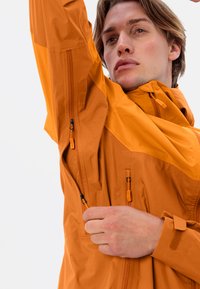 Giacca arancione con zip, colletto alto e polsini regolabili. Presenta zip di ventilazione laterali e sotto le ascelle e una texture del tessuto liscia.