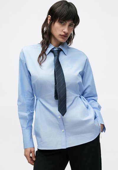 Ανοιχτό μπλε πουκάμισο με κουμπιά και λεπτομέρεια δεσίματος στη μέση, μακριά μανίκια και μπλε ριγέ γραβάτα, συνδυασμένο με μαύρο παντελόνι. Ομοιόμορφη, λεία υφή σε όλο το ύφασμα.