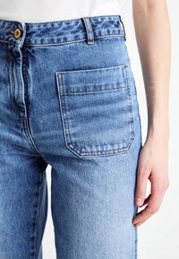 Jeans de mezclilla azul claro con una cintura alta, costuras visibles y un solo bolsillo trasero. La tela está ligeramente deslavada y tiene una textura suave.