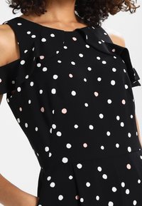 Robe noire avec des pois blancs et roses, présentant un design épaule dénudée et des accents de volants autour du décolleté. Texture de tissu douce.