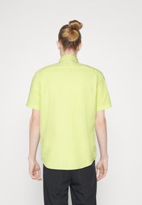 Koszula krótki rękaw w jasnym żółtym kolorze, wykonana z fakturowanej tkaniny i o luźnym kroju. Kołnierz jest stylu bandażowego, a tył ma zaokrąglony dół.