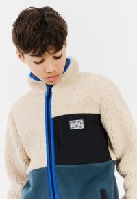 Fleece jacket featuring cream, black, and teal fabric, with a high collar, blue zipper accent, and a logo patch on the chest.