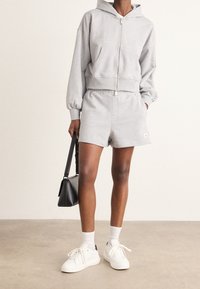 Sudadera gris con cremallera y pantalones cortos con cintura elástica, zapatillas blancas y un bolso negro de hombro. La tela parece suave y tiene un ajuste relajado.