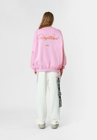 Sudadera rosa de gran tamaño con texto bordado en la parte posterior. Combinada con pantalones blancos que tienen un bolsillo lateral y detalles en cinta negra.