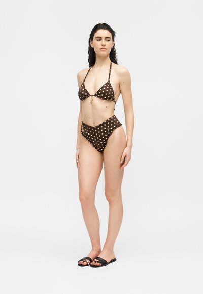 Woman standing in a brown polka dot bikini and black slide sandals, looking to the side against a plain white background.