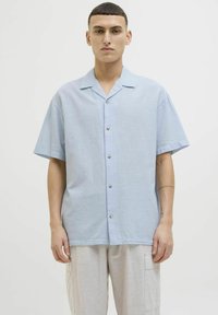 Camisa de manga corta azul claro con un corte relajado, frente abotonado y una textura sutil, combinada con pantalones gris claro.