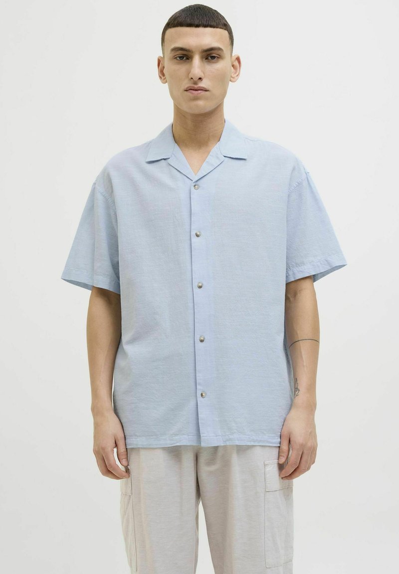 Camisa de manga corta azul claro con un corte relajado, frente abotonado y una textura sutil, combinada con pantalones gris claro.