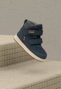 Baskets montantes en suède bleu avec des bandes Velcro, semelle en caoutchouc clair contrastante et col rembourré, exposées sur une surface carrelée texturée.