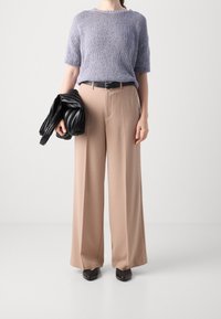 Heller lila Kurzarmstrickpullover, beigefarbene weit geschnittene Hose, schwarze Lederjacke in der Hand, schwarzer Gürtel und schwarze Schuhe.