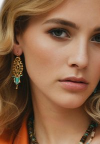 Boucles d'oreilles dorées avec un design géométrique complexe, des accents turquoise et des perles suspendues. La lumière naturelle met en valeur leur texture détaillée.
