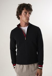 Giacca a zip blu navy in tessuto texture, con accenti a strisce rosse e bianche su colletto e polsini. Indossata sopra una camicia bianca, abbinata a pantaloni chiari.
