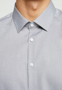 Camicia grigia con bottoni, dal tessuto morbido, dotata di colletto a punta, bottoni bianchi e un motivo tessile delicatamente intrecciato.