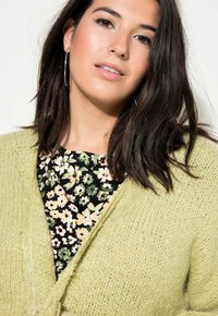 Cárdigan de punto verde claro con superficie texturizada, de frente abierto y bordes deshilachados, sobre una blusa negra con estampado floral que presenta flores rosas.