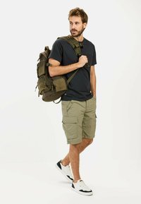 Oliven grøn rygsæk med flere lommer, sort t-shirt og lysegrønne cargo shorts. Hvide sneakers med sorte accenter.