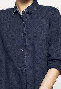 Camicia di jeans blu scuro con chiusura a bottoni, colletto a punta e maniche arrotolate. Presenta una texture sottile e dettagli in bottoni argentati.