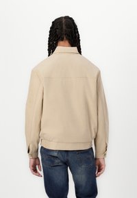 Veste bomber beige avec un col, une taille élastique et des manches longues. Texture douce et design minimaliste, accompagnée d'un jean en denim foncé.