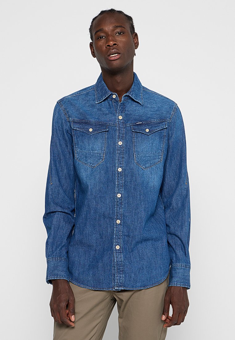 Chemise en denim bleu à manches longues, col boutonné, deux poches poitrine et texture légèrement délavée. Possède des boutons blancs et une coupe ajustée.