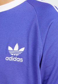 Fioletowa bawełniana koszulka sportowa z białym okrągłym dekoltem, trzema białymi paskami na ramionach oraz haftowanym logo Adidas na piersi.