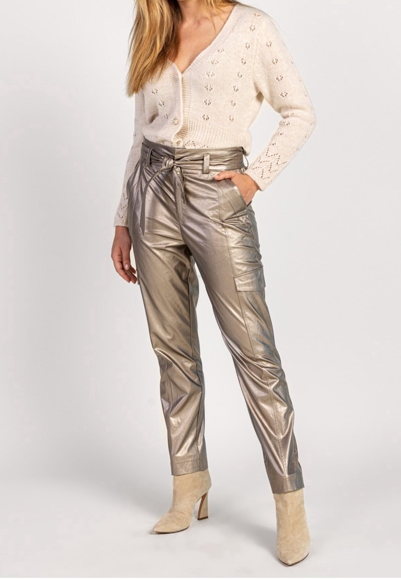 Pantalon doré métallique taille haute avec détail ceinture, associé à un cardigan crème en maille torsadée. Des talons en daim beige complètent la tenue.