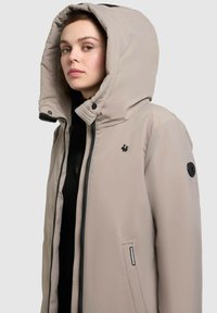 Bežová bunda s kapucňou, ktorá má zips vpredu, čierne akcenty a vyšitý logo. Tkanina sa zdá hladká s miernym leskom.