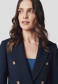 Granatowa marynarka dwurzędowa z złotymi guzikami, dopasowany krój, miękka tekstura, w połączeniu z niebieską bluzką, prezentowana na neutralnym tle.