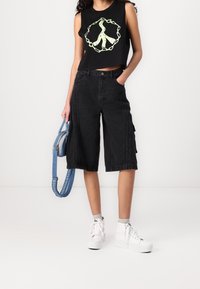 Sort ærmeløs crop top med grafisk design, parret med sorte cargo shorts med sidelommer. Hvide sneakers og blå skuldertaske.