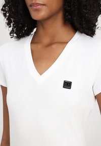 Camiseta blanca de cuello en V, hecha de tela suave, con un parche de logo negro en la parte izquierda del pecho. Mangas cortas y un corte relajado.